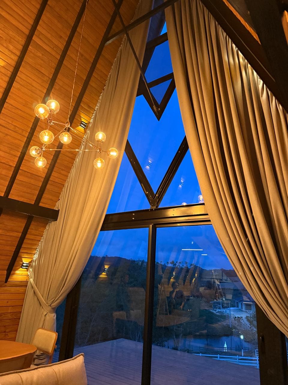 Cortinas claras abertas em uma janela alta de chalé, com teto de madeira e vista ao entardecer