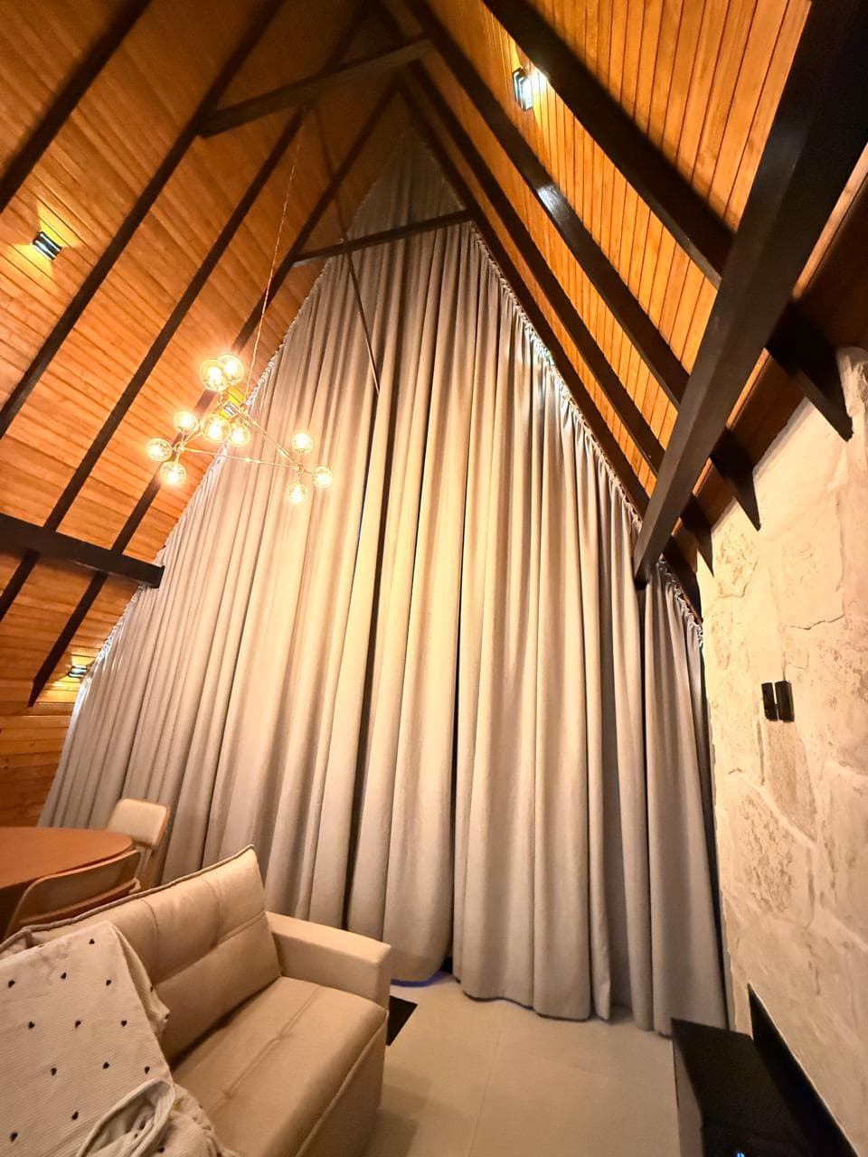 Cortinas claras fechadas cobrindo a janela alta de um chalé com teto de madeira